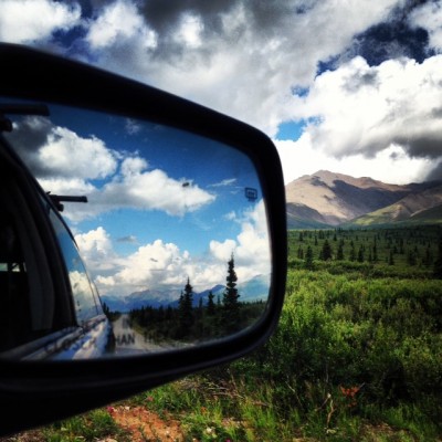Photo was taken looking into the rearview mirror on a car with scenery of Denali National Park ahead and blue skies behind
